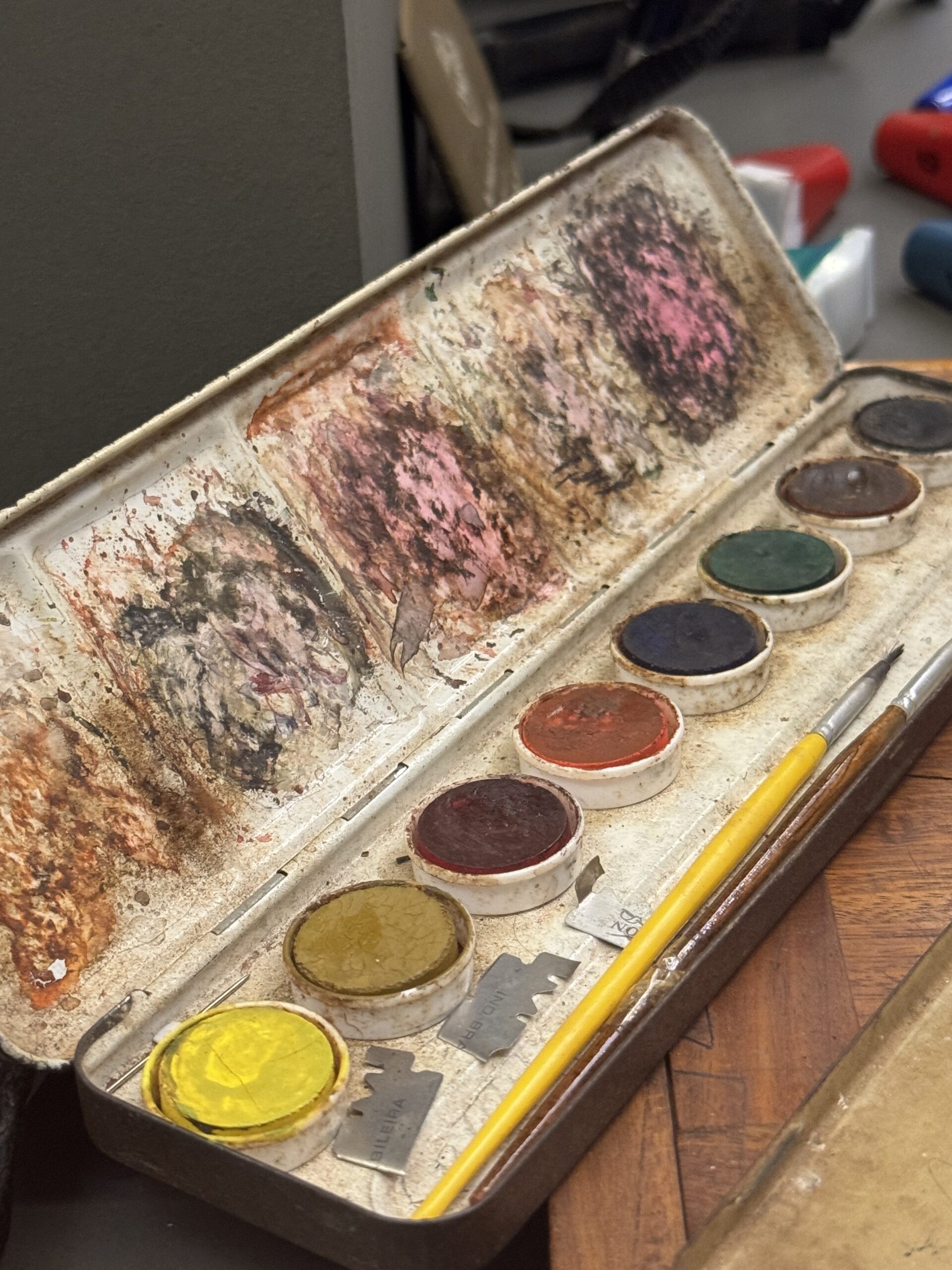 Lata de aquarela usada, com as cubas de mistura cobertas de camadas secas de tinta e dois pincéis largados por cima.